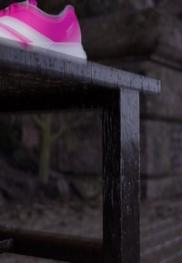 Jasně růžový a bílý teniska odpočívající na mokré tmavé dřevěné lavičce s rozmazaným pozadím kamenné zdi a stromů.