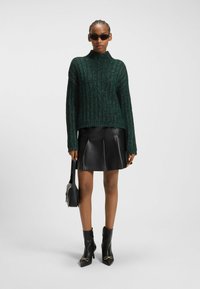 Grön stickad tröja med hög hals, i kombination med en svart plisserad skinnkjol och ankelstövlar. Accenter inkluderar stilrena solglasögon och en handväska.