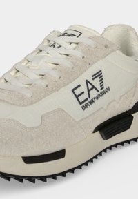 Λευκό και μπεζ sneaker Emporio Armani με υφασμάτινα πάνελ, λευκά κορδόνια, μαύρες λεπτομέρειες στη σόλα και λογότυπο EA7 στο πλευρό.