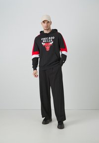 Felpa nera con maniche rosse e bianche, con testo e logo "Chicago Bulls". Pantaloni larghi neri e berretto beige. Scarpe nere.