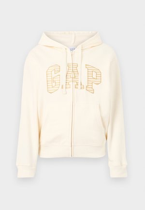 Hoodie zippé de couleur crème avec poches avant, cordons de serrage et logo "GAP" en lettres rayées dorées sur la poitrine.