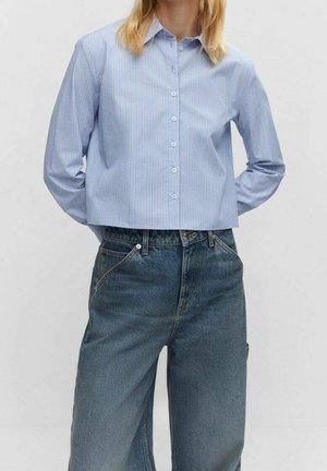 Frau trägt ein hellblau gestreiftes Hemd mit langen Ärmeln, das hinten eingesteckt ist, und eine hoch taillierte, lockere blaue Jeans.