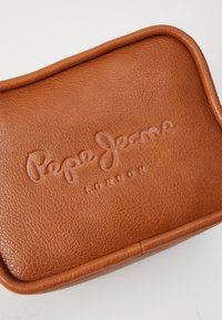 Ruda oda toilečių kuprinė su embosiuotu "Pepe Jeans London" logotipu. Suapvalinti kampai, tekstūruotas paviršius, užtrauktuko uždarymas ir jokie matomi raštai.
