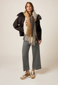 Braun-grauer Farbverlaufsschal, übergroße dunkle Jacke, cremefarbenes Grafik-T-Shirt, graue Hosen mit weitem Bein und braun-beige Sneakers mit weißen Akzenten.