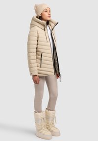 Béžová nadýchaná bunda s vysokým golierom, bočnými zipsami a umelým kožušinovým podšívkom, skombinovaná so svetlobežovými legínami a zimnými čižmami cez kolená.