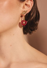 Náušnice v zlatej farbe s dizajnom slučky a príveskom v tvare červenej jablka so zlatým stredom a čiernymi detailmi, pripnuté na uchu.