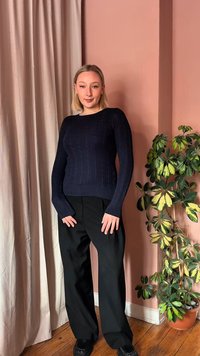 Mladá žena v námorníckom modrom svetri a čiernych nohaviciach stojí vo vnútri vedľa zeleno-žltej kvetináčovej rastliny na pozadí broskyňovej steny.