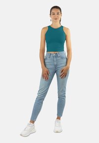 Frau steht in einem türkisfarbenen ärmellosen Crop-Top, hellblauen hochgeschnittenen Jeans, weißen Sneakern, mit nach hinten gebundenem Haar und neutralem Ausdruck.