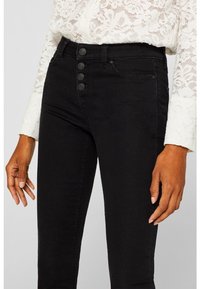 Pantalones vaqueros negros de cintura alta con cierre de cinco botones, con una textura suave y un diseño minimalista. Combinado con una blusa de encaje blanca.
