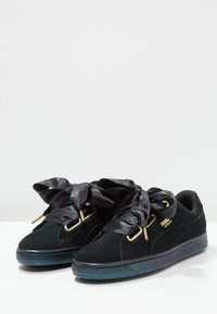 Baskets en daim noir Puma avec des lacets en ruban satin épais et des œillets dorés, présentées côte à côte sur une surface blanche.