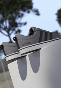 Zapatillas Adidas grises con rayas negras descansando sobre un saliente blanco al aire libre bajo un cielo azul claro con árboles desenfocados al fondo.