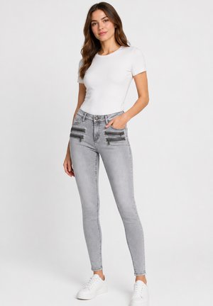 Kvinde står iført en tætsiddende hvid kortærmet top, lysegrå skinny jeans med fire lynlåse foran og hvide sneakers.