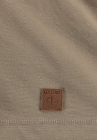 Tela en color beige con una textura suave, presenta un dobladillo cosido y una etiqueta de cuero marrón con la inscripción "KEY LARGO" acompañada de un diseño de logo.
