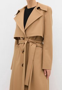 Personne portant un trench-coat camel ceinturé avec de grands revers et des boutons en écaille sur un haut noir.