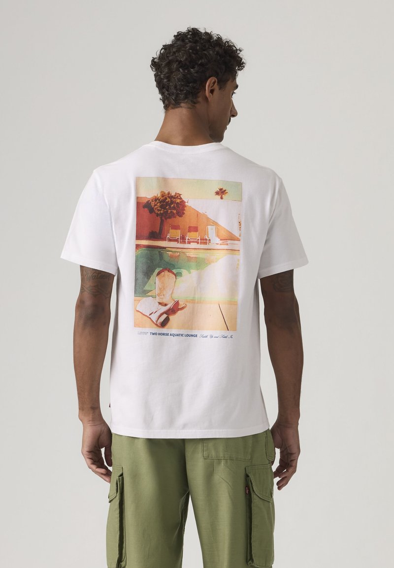 Mann mit lockigem Haar, der ein weißes T-Shirt mit farbenfrohem Pool-Motiv und grüne Cargo-Hosen trägt, steht vor einem schlichten Hintergrund.