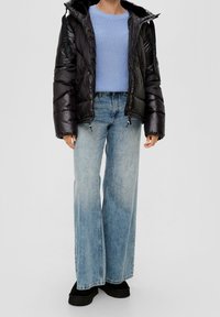 Schwarze Daunenjacke mit Kapuze, hellblaues Strickoberteil und weit geschnittene blaue Jeans. Schwarze Plateauschuhe vervollständigen das Outfit.