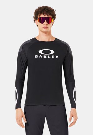 Homme portant un t-shirt noir à manches longues Oakley et un pantalon assorti avec de grandes lunettes de soleil de sport rouges, debout devant un fond uni.