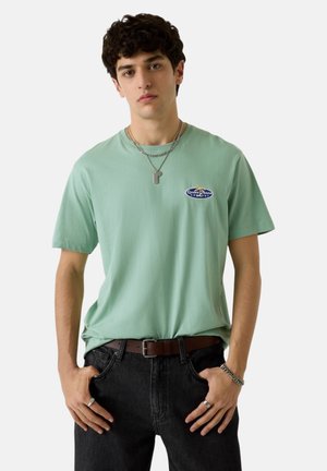Ragazzo giovane con capelli ricci scuri che indossa una T-shirt verde chiaro con logo, jeans scuri, cintura marrone, collana d'argento, anelli e braccialetto, in piedi con le mani nelle tasche.