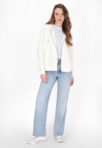 Světle krémová mikina na zip s šňůrkou, přes bílou a modrou pruhovanou crop top. Volné světle modré džíny a bílé tenisky.