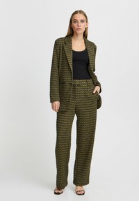 Ensemble coordonné comprenant un blazer à motifs texturés vert et noir et un pantalon large, associé à un débardeur noir et des talons.