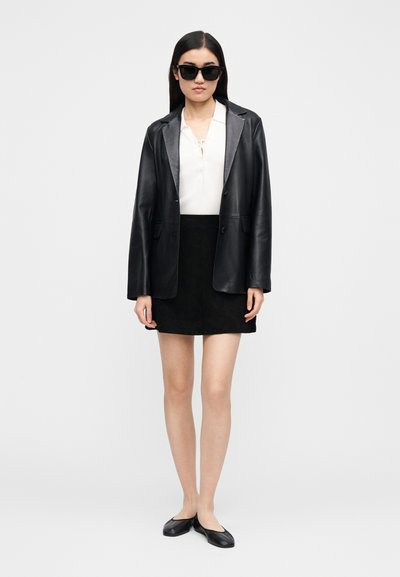 Black leather blazer over a white top with a tie detail, paired with a black suede mini skirt and black flat shoes, against a white background.