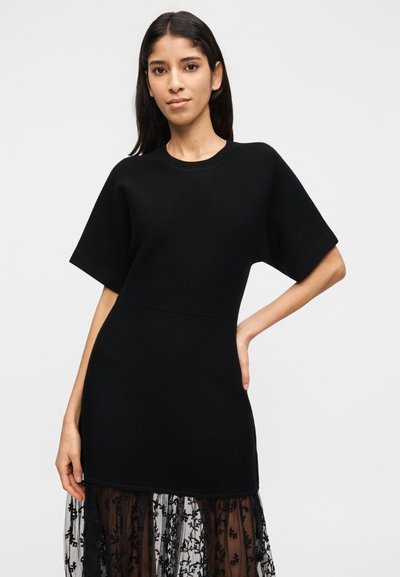 Γυναίκα με μακριά σκουρόχρωμα μαλλιά φορά ένα μαύρο κοντομάνικο φόρεμα πάνω από διαφανές δαντελένιο φούστα, ποζάροντας με το ένα χέρι στη μέση μπροστά σε απλό φόντο.