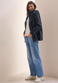 Marineblå vattentät jacka med huva, knappstängning och skräddarsydd passform; ihop med ljusblå jeans och vit-gröna sneakers. Slät textur.