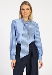 Blonde vrouw in een blauw gestreepte blouse met een versierde kraag en strik, gecombineerd met hooggesneden donkerblauwe broek.