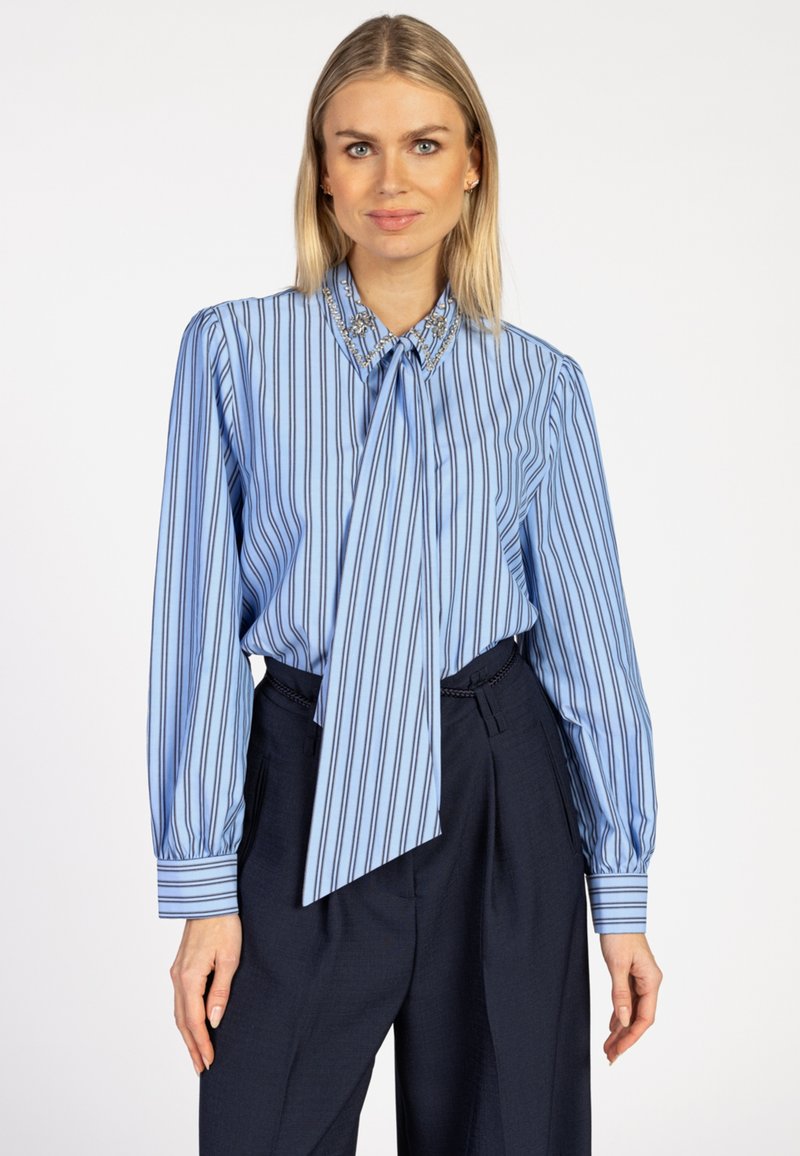 Blonde vrouw in een blauw gestreepte blouse met een versierde kraag en strik, gecombineerd met hooggesneden donkerblauwe broek.