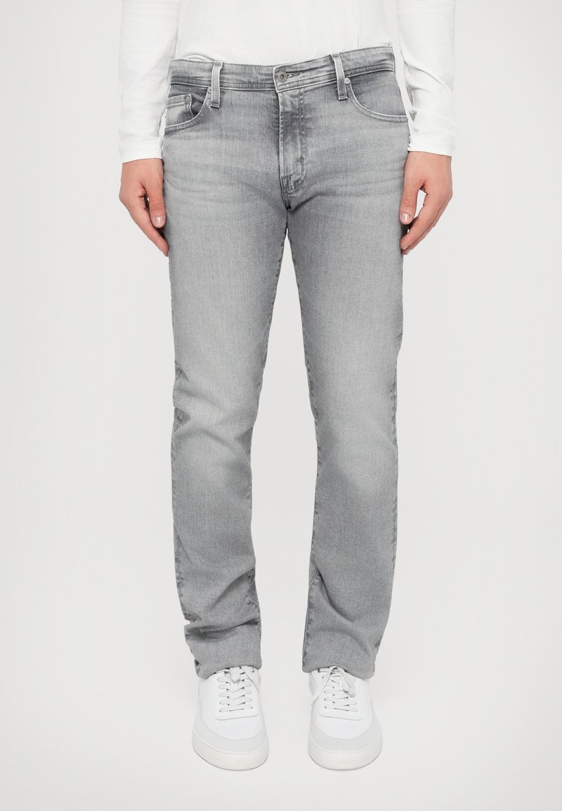 Graue Jeans aus Denim mit geradem Schnitt, zwei Fronttaschen, leichten Abnutzungen und einem Reißverschluss, kombiniert mit weißen Sneakers.