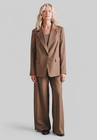 Brązowy garnitur na miarę z dopasowaną marynarką zapinan�ą na jeden guzik, dwoma kieszeniami z przodu oraz szerokimi spodniami, zestawiony z prostym topem.