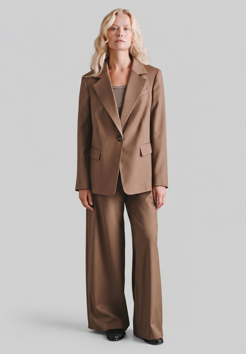 Brązowy garnitur na miarę z dopasowaną marynarką zapinaną na jeden guzik, dwoma kieszeniami z przodu oraz szerokimi spodniami, zestawiony z prostym topem.