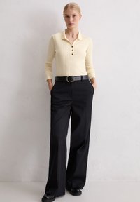 Frau, die gegen eine weiße Wand steht, trägt ein cremefarbenes, geripptes Langarmoberteil mit schwarzen Knöpfen, schwarze weite Hosen sowie einen schwarzen Gürtel und schwarze Schuhe.