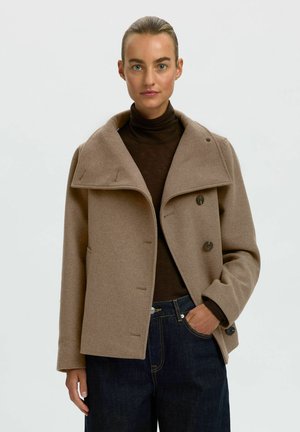 Femme aux cheveux tirés en arrière porte un manteau beige à double boutonnage sur un col roulé marron foncé et un jean bleu foncé, main dans la poche, fond neutre.