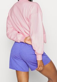 Sweat léger rose à épaules tombantes associé à un short violet, tous deux en tissu doux, offrant une coupe décontractée.