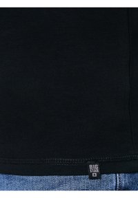 Schwarzer Stoff mit sichtbaren Nähten und einem kleinen schwarzen Etikett mit der Aufschrift "BIG STAR" über blauen Denim-Jeans.