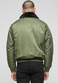 Alyvuogių žalios spalvos bombonešio stiliaus striukė su tekstūruotu juodos spalvos dirbtinės kailinės apykakle, ribotomis rankovėmis ir juosmeniu, lygiu išoriniu paviršiumi ir paminkštu dizainu.