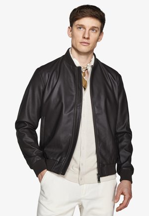 Młody mężczyzna w czarnej skórzanej kurtce typu bomber, beżowej koszuli zapinanej na guziki, lekkim szaliku i białych spodniach, stoi z jedną ręką w kieszeni.