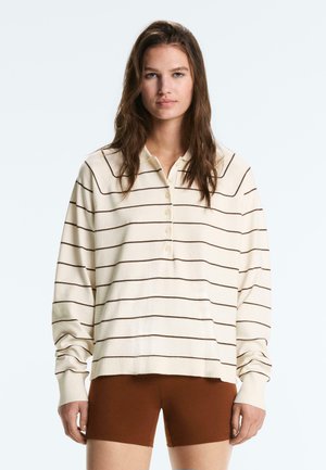 Žena v krémovom poloroláku s dlhými rukávmi s tenkými hnedými horizontálnymi pruhmi a priliehavých hnedých kraťasoch stojaca pred jednoduchým pozadím.