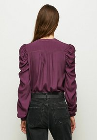 Vista posteriore di una blusa a maniche lunghe di colore viola con maniche arricciate e un design a yoke smock, abbinata a jeans neri a vita alta.