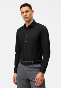Chemise noire à manches longues avec un col classique, fermeture à boutons, et texture lisse, portée avec un pantalon gris à motifs et une ceinture noire.