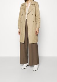 Béžový trenčkot s dvouřadým zapínáním, velkým límcem a dlouhými rukávy, spojený s hnědými širokými kalhotami a bílými kotníčkovými botami.