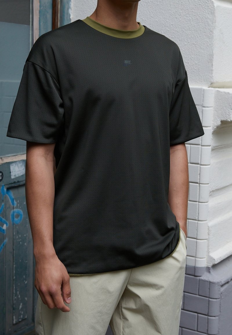 Camiseta negra de manga corta hecha de un material texturizado con un cuello verde y un sutil logo de Nike en el pecho. Combinada con pantalones de color claro.