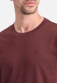 Burgundia värvi puuvillane lühikeste varrukatega T-särk, millel on rinnal väike tikandiga logo ja sile, tekstureeritud viimistlus.