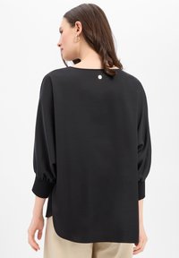 Czarna bluzka o luźnym kroju, z długimi bufkami na rękawach i zaokrąglonym dekoltem. Z tyłu mały guzik jako detal. Gładka faktura.