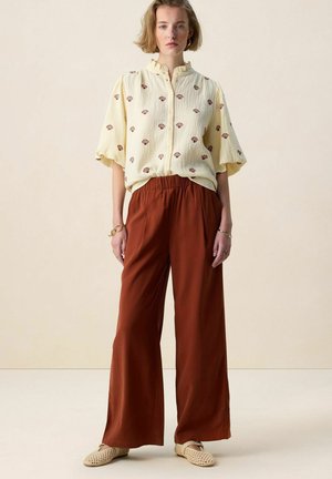 Femme portant un chemisier ample crème à imprimés coquillages, un pantalon large couleur rouille et des chaussures tissées beige, posant devant un fond clair uni.