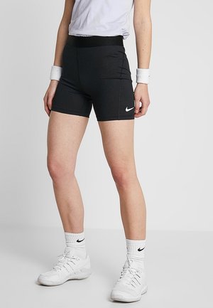 Kobieta ubrana w czarne sportowe szorty Nike, białe skarpetki Nike, białe trampki, białe opaski na nadgarstek i białą koszulkę, stojąca na jednolitym tle.