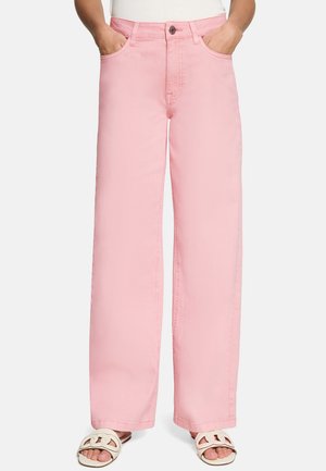 Személy magas derekú, bő szárú rózsaszín nadrágot visel, kezét zsebében tartja, fehér papucsokkal párosítva.