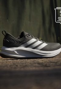 Chaussure de running Adidas noire et blanche avec trois bandes blanches et semelle épaisse rembourrée portant l'inscription "DREAMSTRIKE" sur un trottoir extérieur.