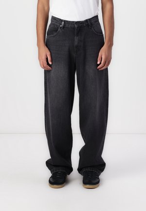 Personne portant un jean en denim noir ample avec des jambes larges et des baskets noires avec des semelles en gomme, debout devant un fond blanc uni.